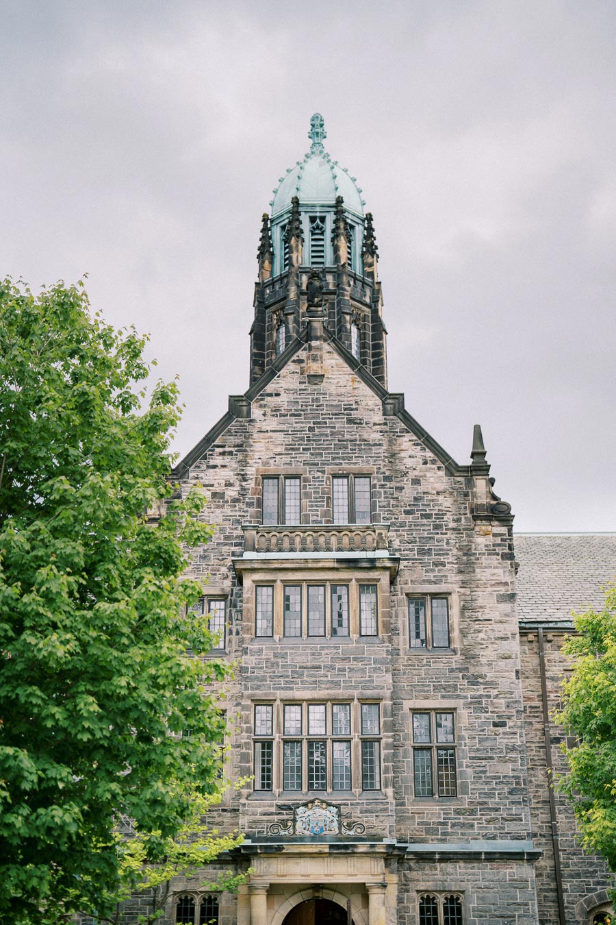 Historic stone building with ornate tower and lush green trees in front, under a cloudy sky
