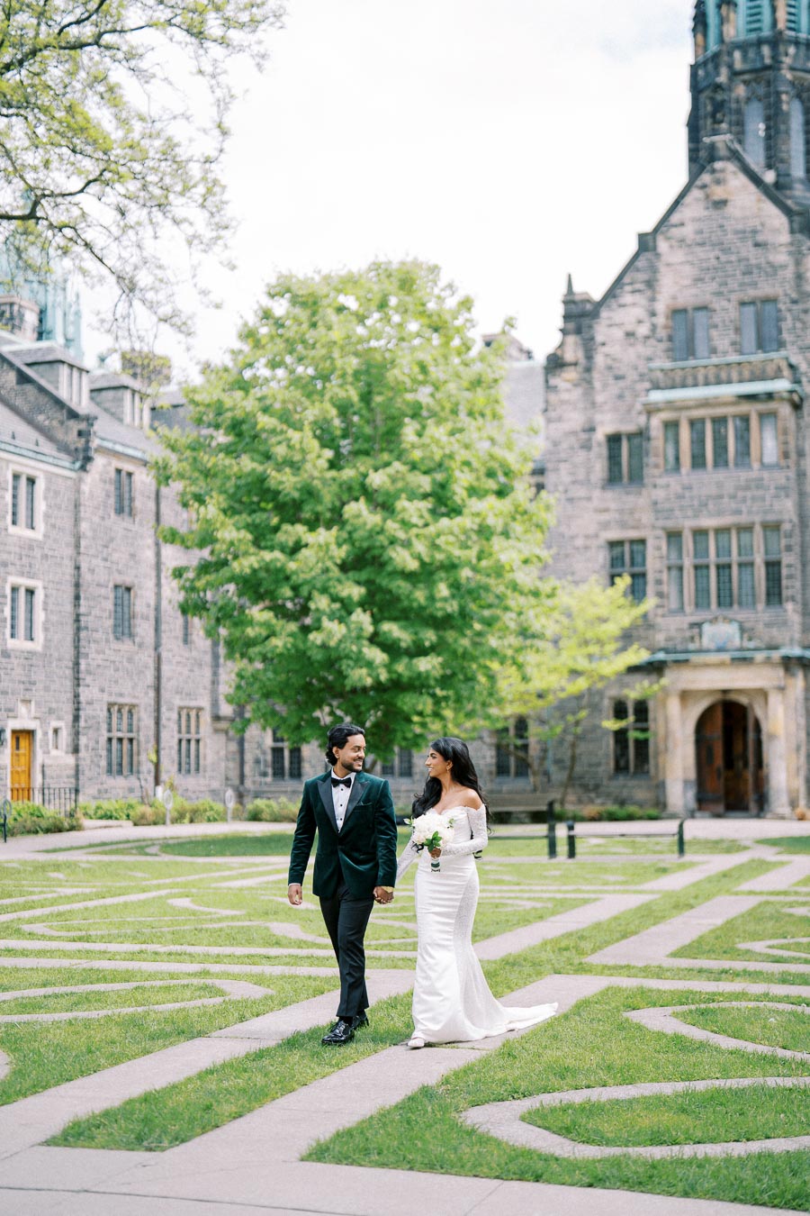 A bride and groom walking hand in hand through a picturesque courtyard with intricate stone pathways and historic architecture in the background, surrounded by lush greenery.