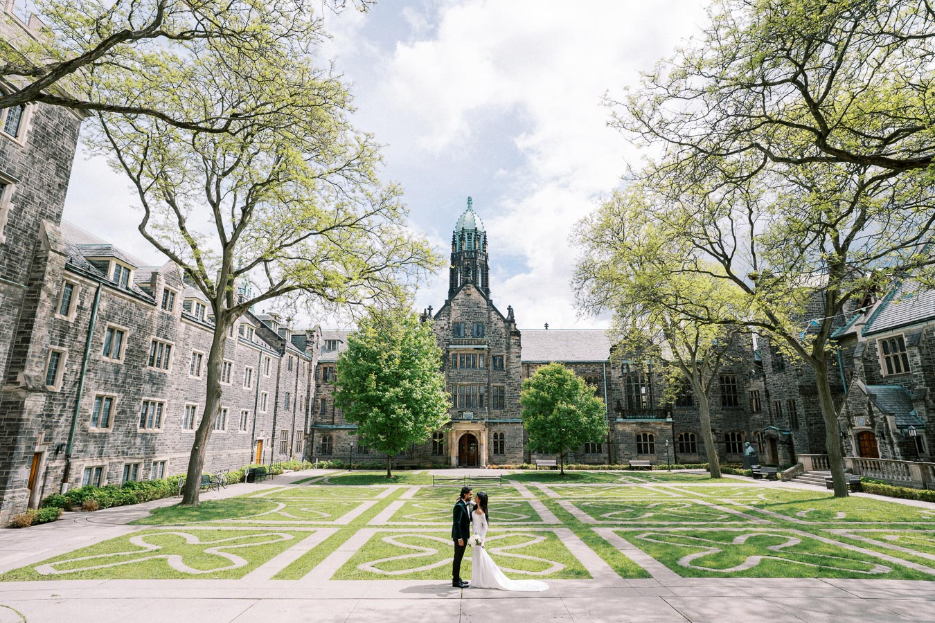A couple in wedding attire stands in a beautifully landscaped courtyard with intricate stone pathways and lush green trees, set against the backdrop of a historic, Gothic-style building, under a partly cloudy sky.