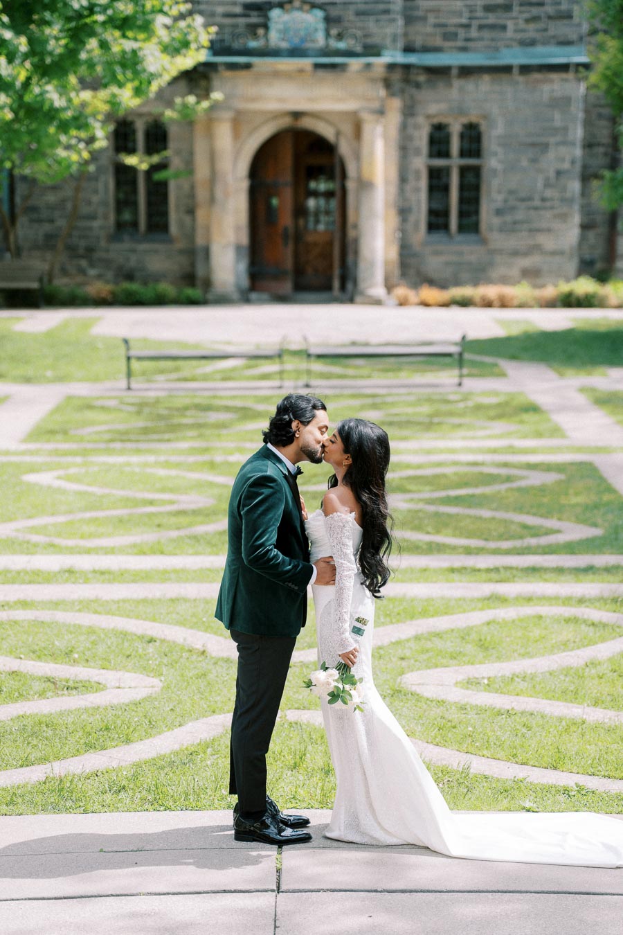 A couple shares a romantic kiss in a beautifully landscaped garden, surrounded by greenery and historic architecture. The bride wears an elegant white gown, holding a bouquet, while the groom is dressed in a dark suit.