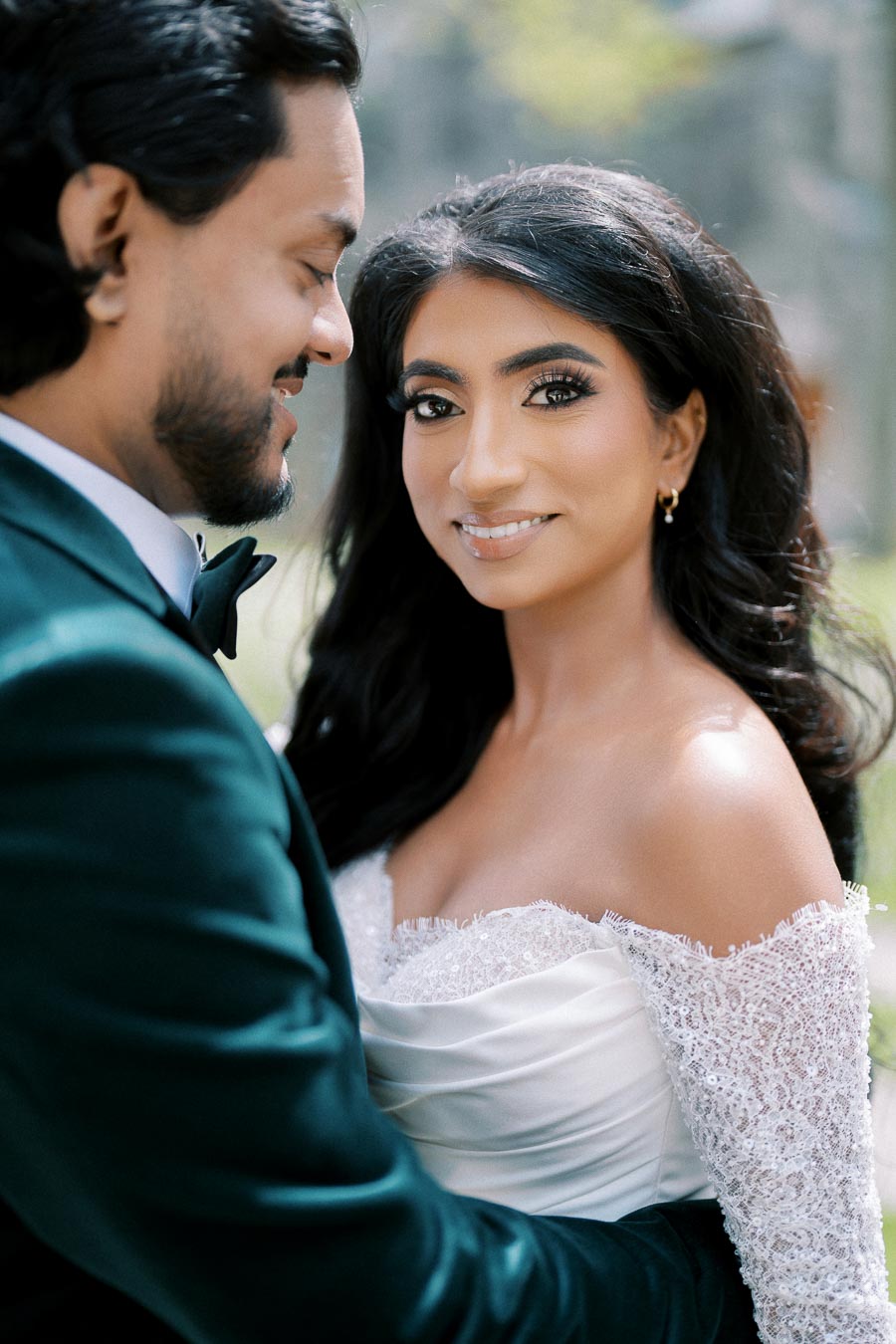 Elegant bride and groom embracing in a romantic outdoor setting, the bride in a lace off-the-shoulder gown and the groom in a dark suit, capturing a joyful wedding moment.