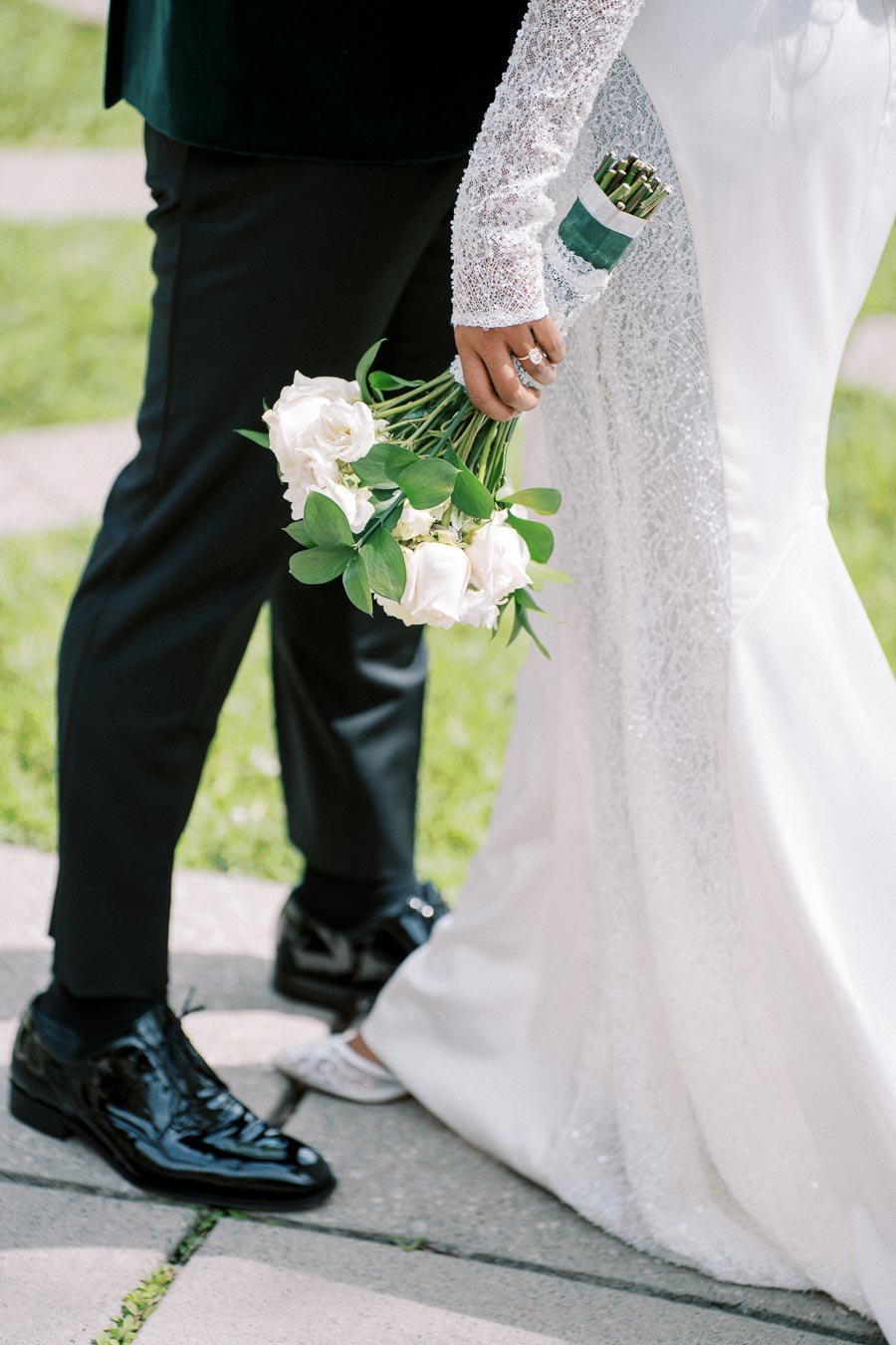 A bride in a white wedding dress holding a bouquet of white roses, standing next to the groom in a black suit and shiny black shoes, with a green grassy background.