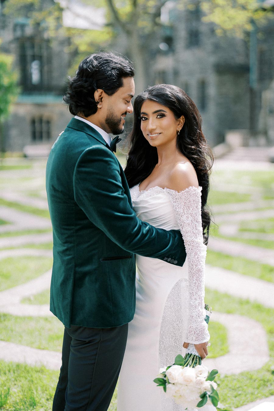 A newlywed couple embracing outdoors in a garden, with the bride in a white gown holding a bouquet of white flowers and the groom in a dark green suit, both smiling and surrounded by lush greenery.