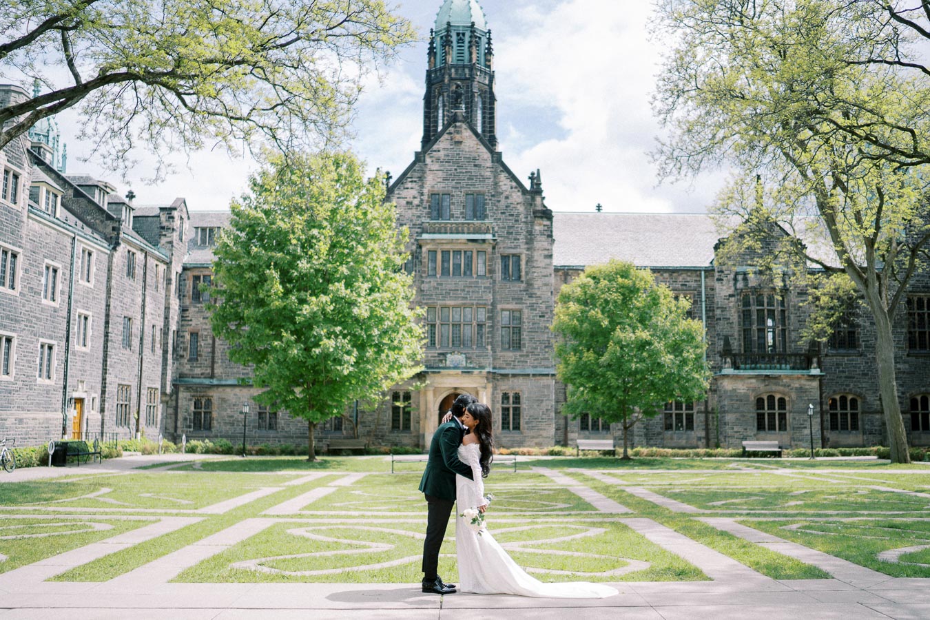 A bride and groom embrace on their wedding day in front of a historic stone university building, surrounded by lush green trees and manicured lawns.
