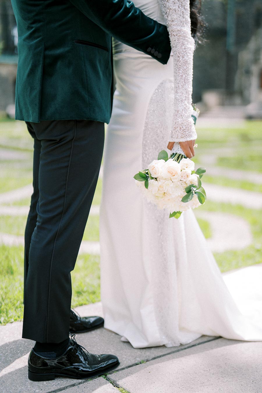 A bride and groom stand closely together on a grassy path, with the bride holding a bouquet of white roses. The groom is in a dark green suit and the bride in a long-sleeved white gown.