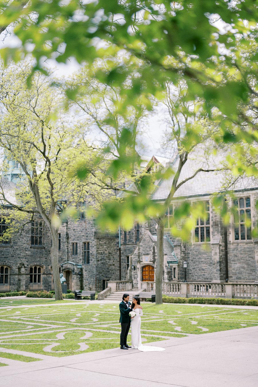 A couple standing in front of a historic stone building, surrounded by lush trees and manicured lawns, during a wedding photoshoot.