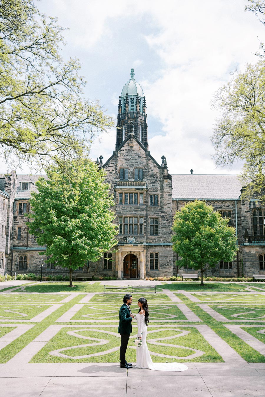 Wedding couple standing in front of historic stone building surrounded by green trees and manicured lawns.