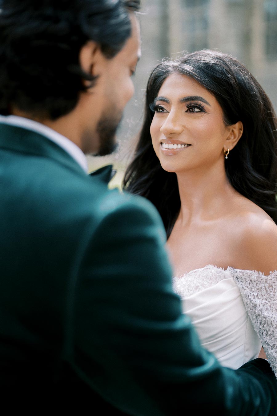 A bride in an elegant white gown smiling at her groom, who is wearing a dark suit, during their outdoor wedding ceremony.