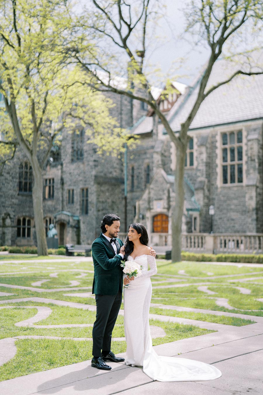 Wedded couple standing in lush garden outside historic stone building, groom in green velvet jacket, bride in elegant white gown holding bouquet, springtime wedding scene.