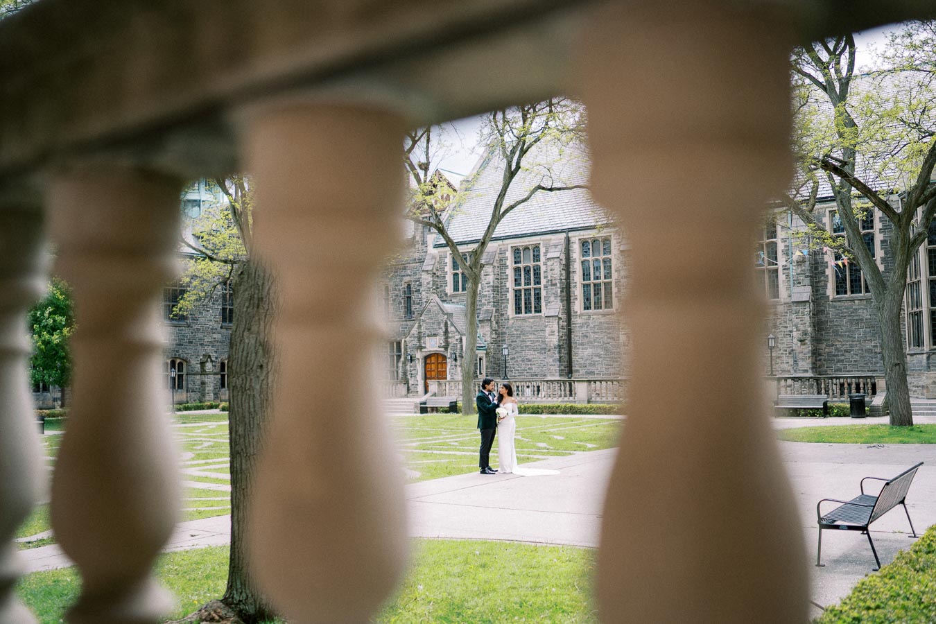 A romantic couple shares a tender moment in front of a historic stone building, framed artistically by foreground architectural columns in a serene courtyard setting.