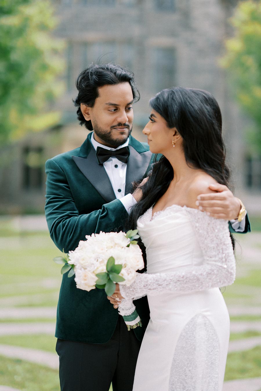 Elegant wedding couple embracing outdoors, groom in a dark green velvet tuxedo and bride in a white lace gown holding a bouquet of white flowers.