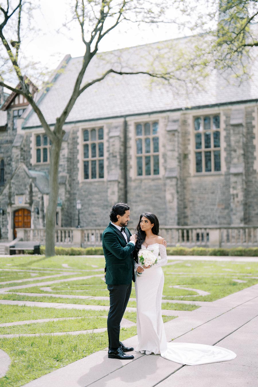 A bride and groom standing together in front of a historic stone church, with the bride wearing an elegant white gown and holding a bouquet, while the groom is dressed in a dark suit.