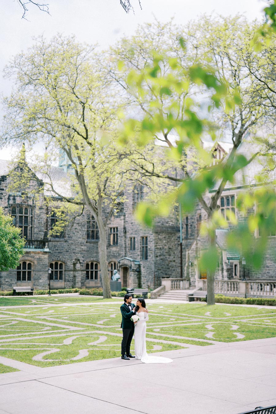 A couple stands in a courtyard with a historic stone building, surrounded by trees and lush greenery, capturing a romantic, outdoor wedding moment.
