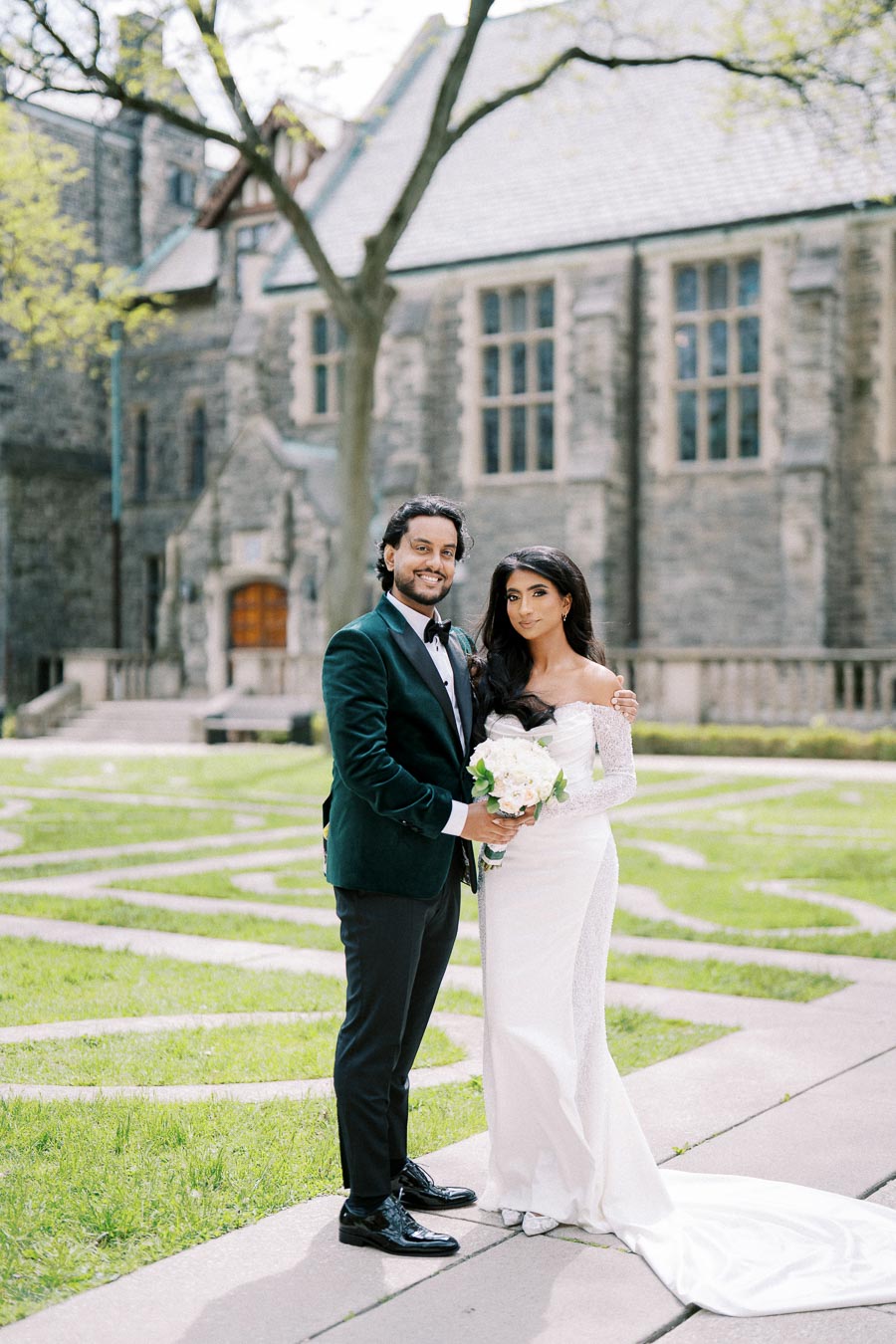 A bride in a long white wedding dress and groom in a green velvet tuxedo pose outdoors in front of a historic stone building, surrounded by lush greenery.