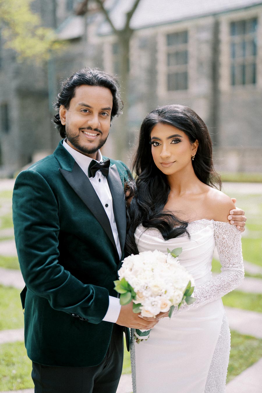 A happy couple posing for a wedding photo, with the groom in a green velvet tuxedo holding a bouquet, and the bride in an elegant white gown, standing outdoors in a garden setting.