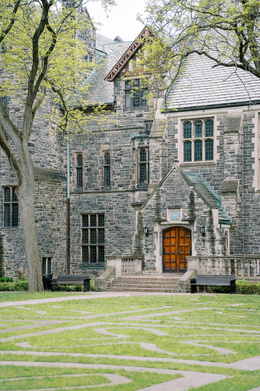 Historic stone university building with arched wooden door, surrounded by lush green grass and bare trees on a bright day.
