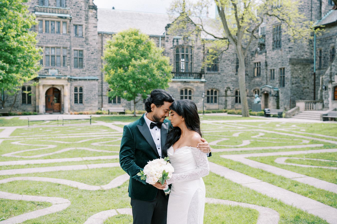 A bride and groom embracing in front of a historic stone building with intricate pathways on a green lawn, creating a romantic wedding scene.