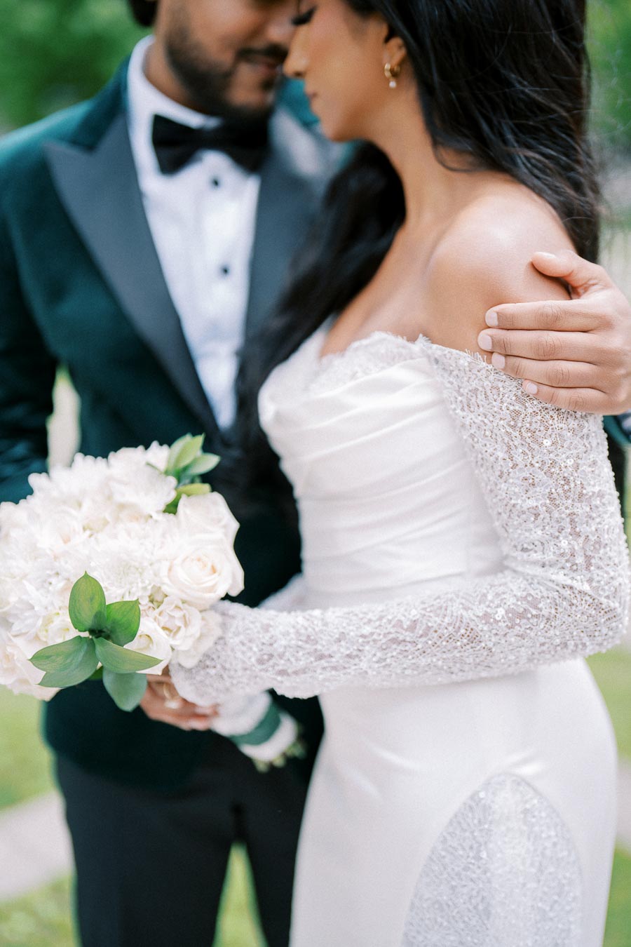 A close-up of a bride and groom on their wedding day. The bride is wearing a white off-the-shoulder gown with intricate lace sleeves and is holding a bouquet of white flowers and greenery. The groom is dressed in a dark suit with a bow tie, gently embracing the bride. They are outdoors, surrounded by greenery, exuding romance and