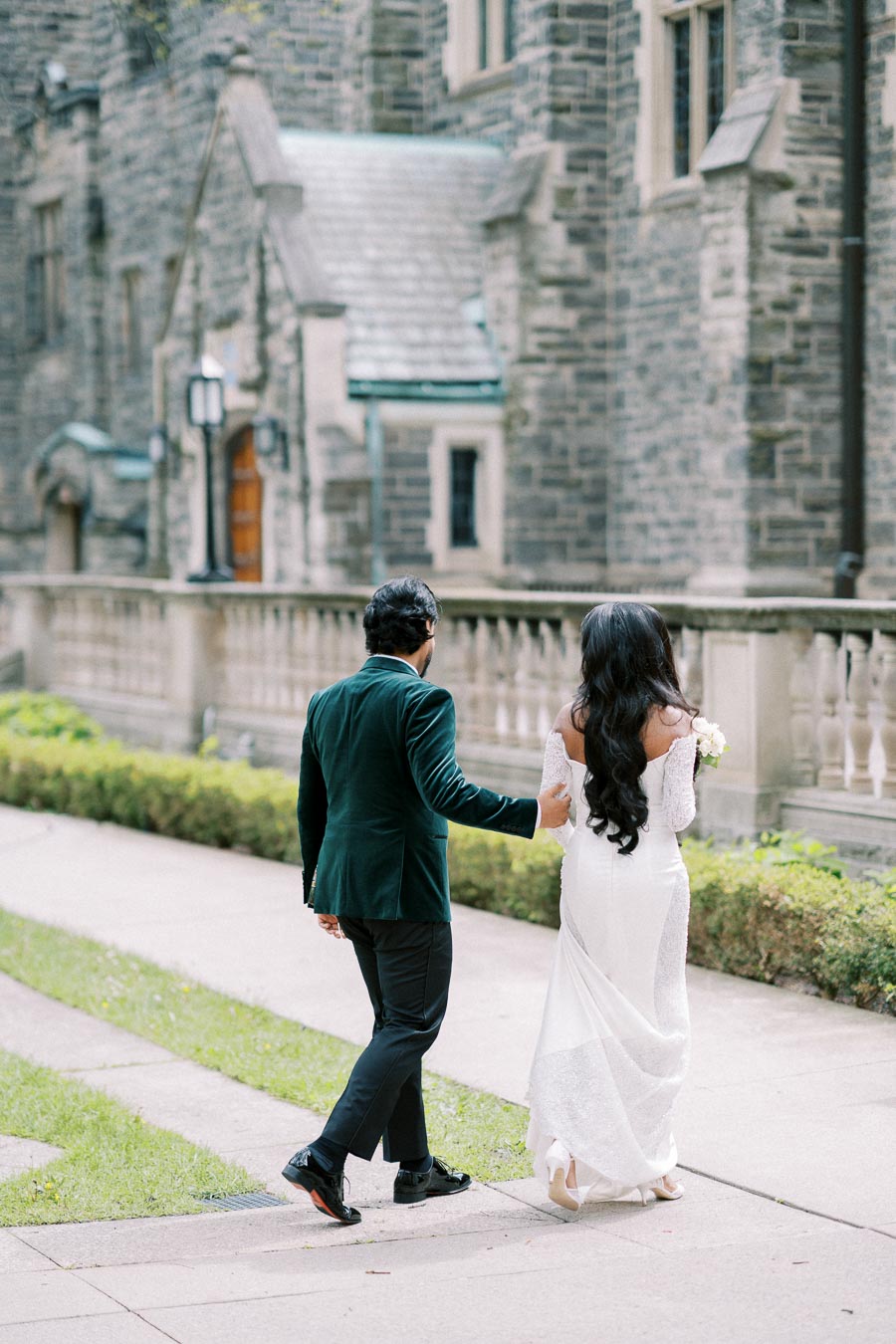 Elegant couple walking outside a historic stone building, with the woman in a white dress holding a bouquet, creating a romantic scene.