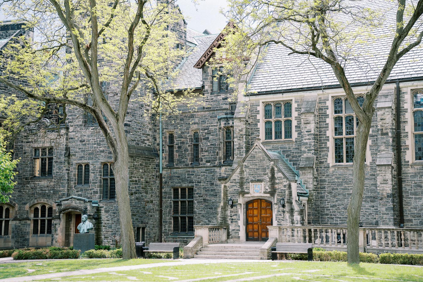Historic stone building with arched windows and wooden door, surrounded by trees and greenery on a bright day.