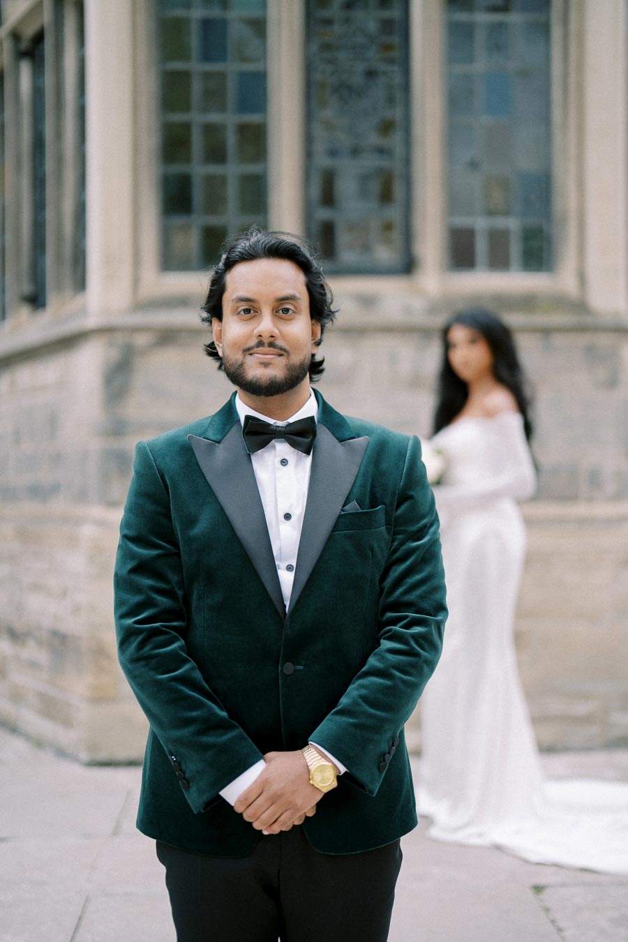 Young man in a velvet tuxedo standing confidently in front of a blurred bride in a white dress, set against a historic stone building with stained glass windows, capturing a wedding moment.