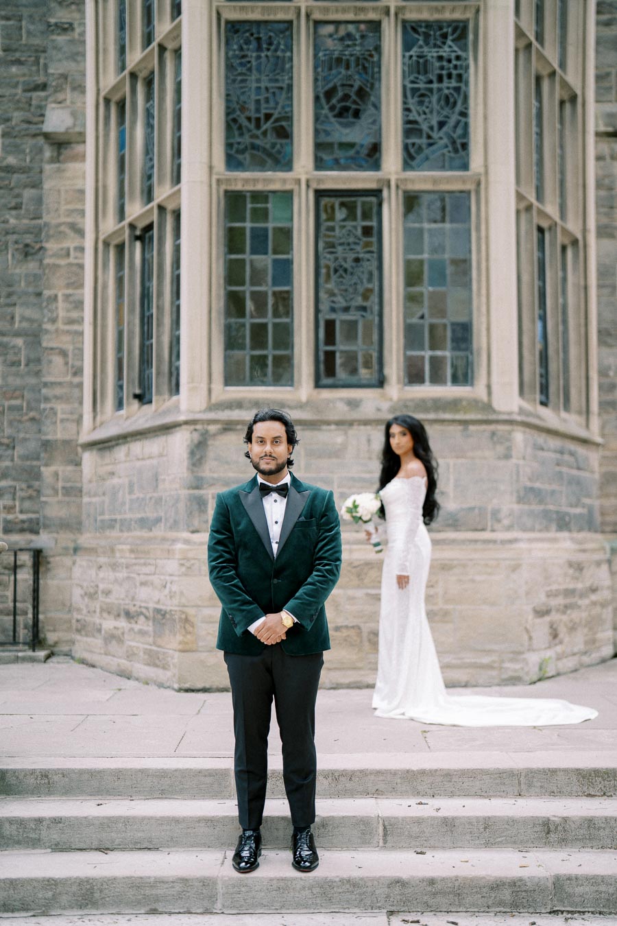 A groom in a dark green velvet suit stands on stone steps, with a bride in a white gown holding a bouquet in the background. The couple is positioned in front of a building with intricate stained glass windows.