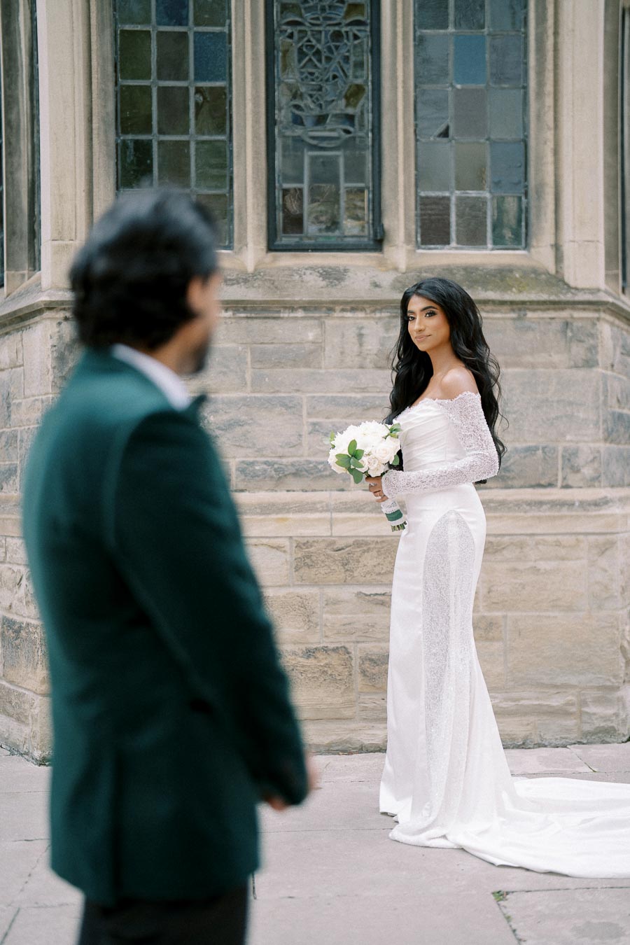 Bride in elegant white gown holding a bouquet, standing outside a stone building with stained glass windows, while a man in a suit looks on.