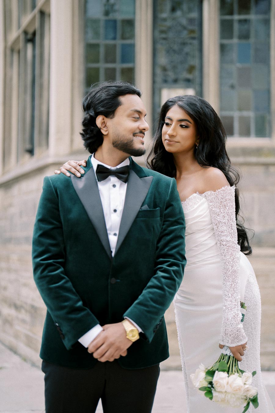 Elegant couple in formal attire with groom in green velvet tuxedo and bride in lace off-shoulder wedding dress holding bouquet, posing outdoors by stone building.