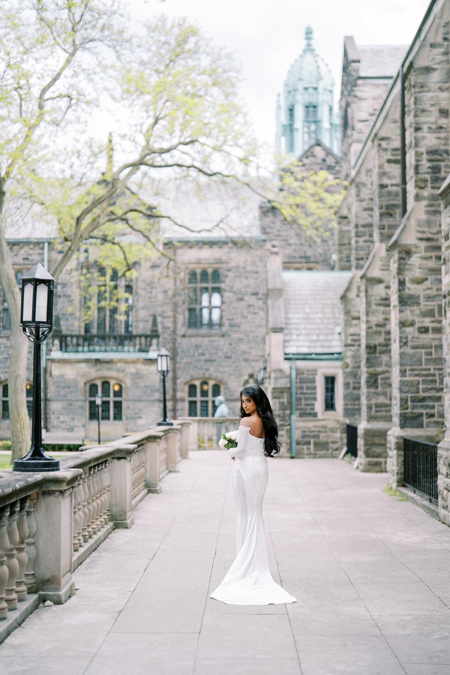 A bride in a white wedding dress holding a bouquet poses on a historic stone pathway, surrounded by gothic architecture and trees with fresh spring leaves.