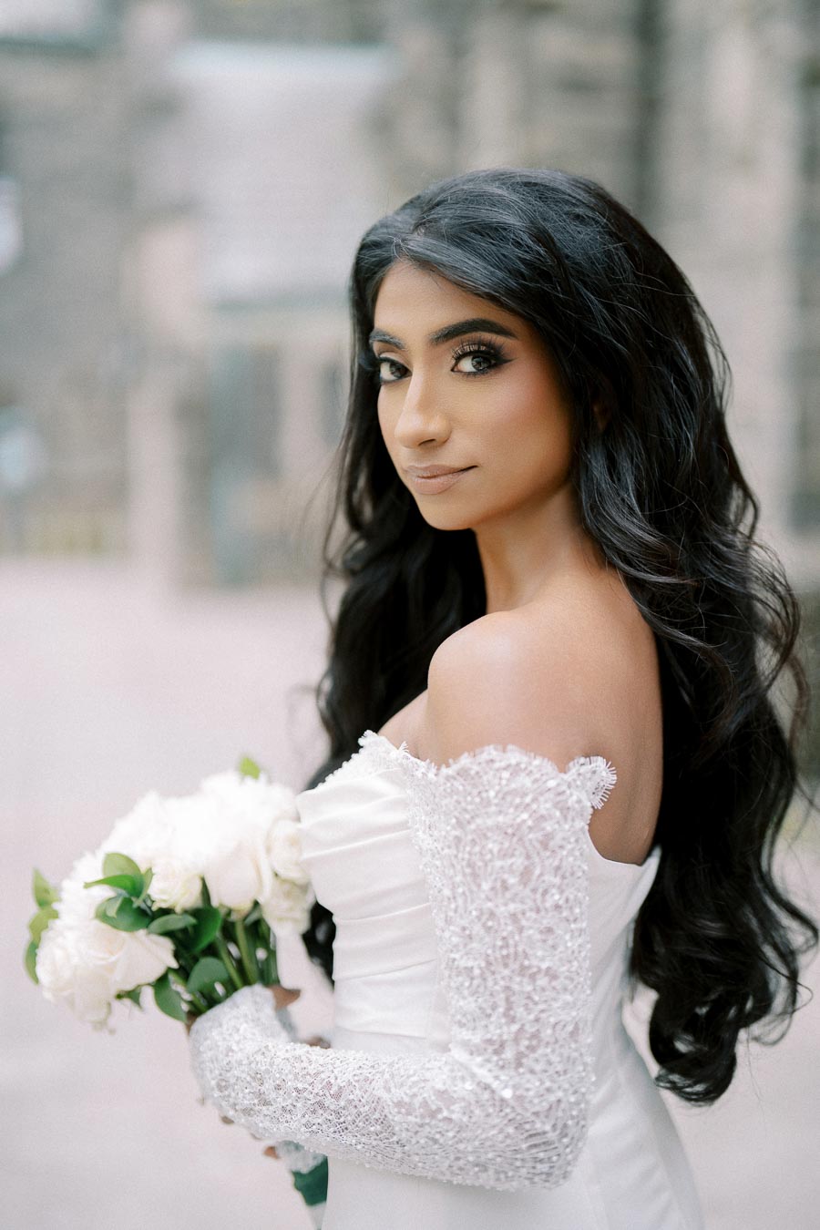 A bride wearing an elegant off-shoulder white wedding dress with lace sleeves, holding a bouquet of white roses, posing outdoors with a soft background.