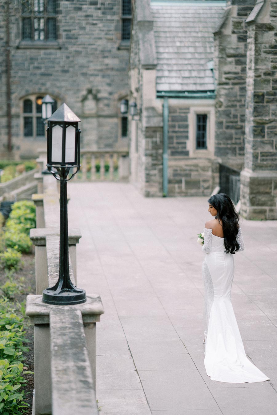 Bride in elegant white gown holding a bouquet, walking along a stone path beside historic architecture, with a vintage lamp post in the foreground.