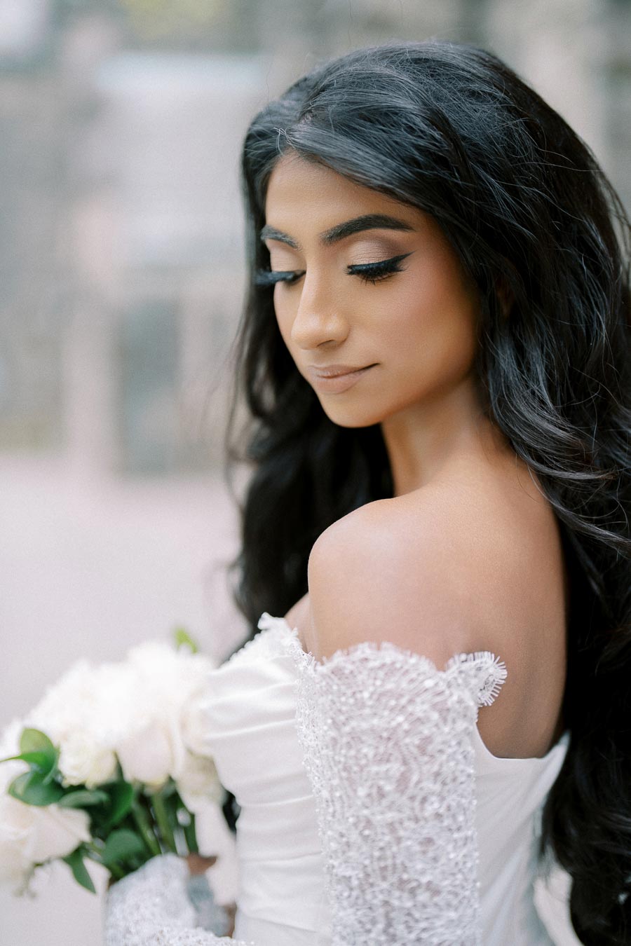 Elegant bride in an embellished white wedding dress holding a bouquet of white roses, with soft romantic makeup and flowing dark hair, set against a blurred outdoor background.