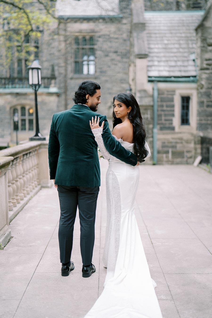 Romantic couple in elegant wedding attire pose on a historic stone walkway, with vintage architecture in the background.
