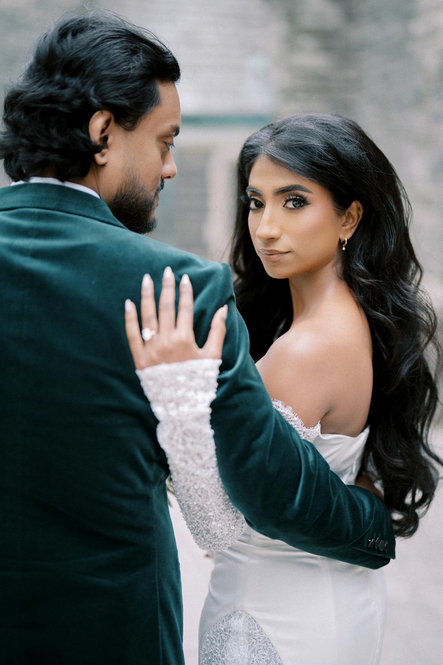 Elegant wedding couple gazing into camera, bride in white off-shoulder gown with lace details and groom in dark green velvet suit, showcasing engagement ring in intimate pose.