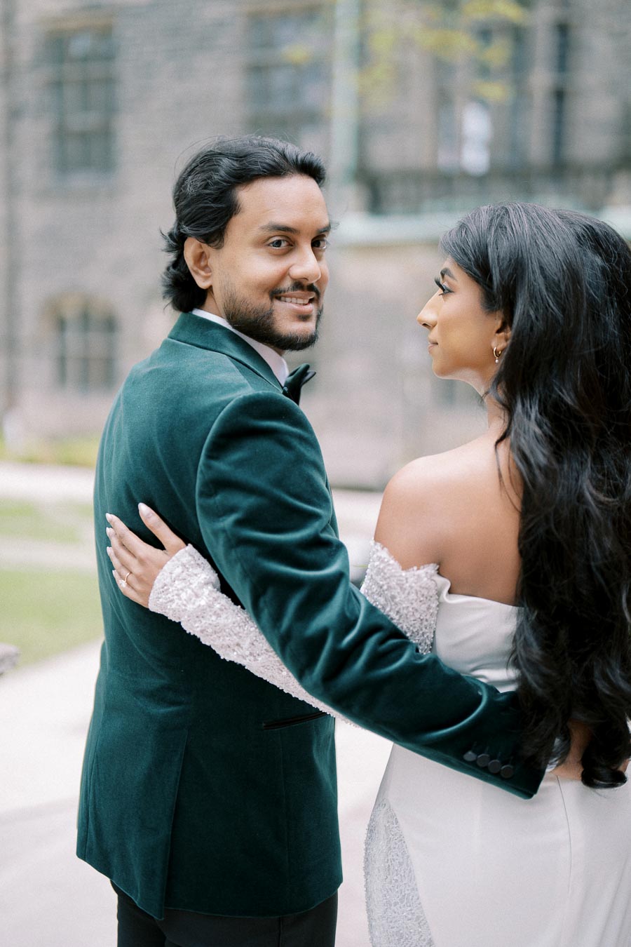A couple in formal attire embracing outdoors, the man wearing a green velvet suit and the woman in a white dress with lace sleeves, with a historic stone building in the background.