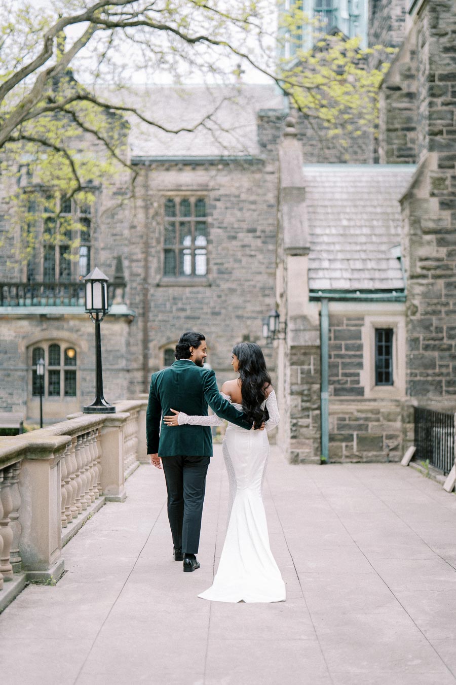 A couple walking arm in arm along a stone pathway, surrounded by historic architecture and lush greenery, wearing elegant formal attire—ideal image for wedding or engagement themes.