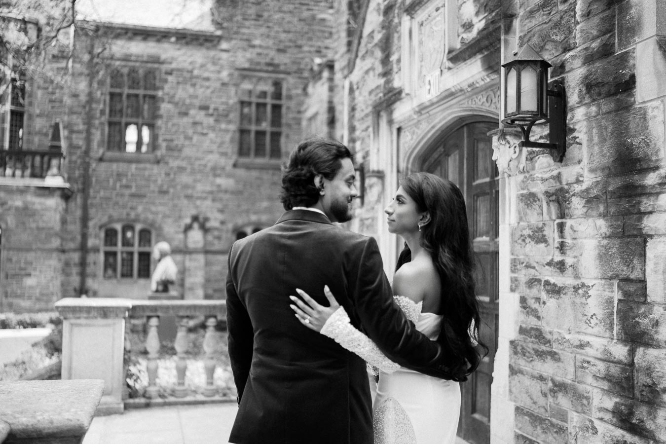 Black and white photo of a couple embracing outside a historic stone building, with the woman in a white dress and the man in a dark suit, capturing a romantic moment.