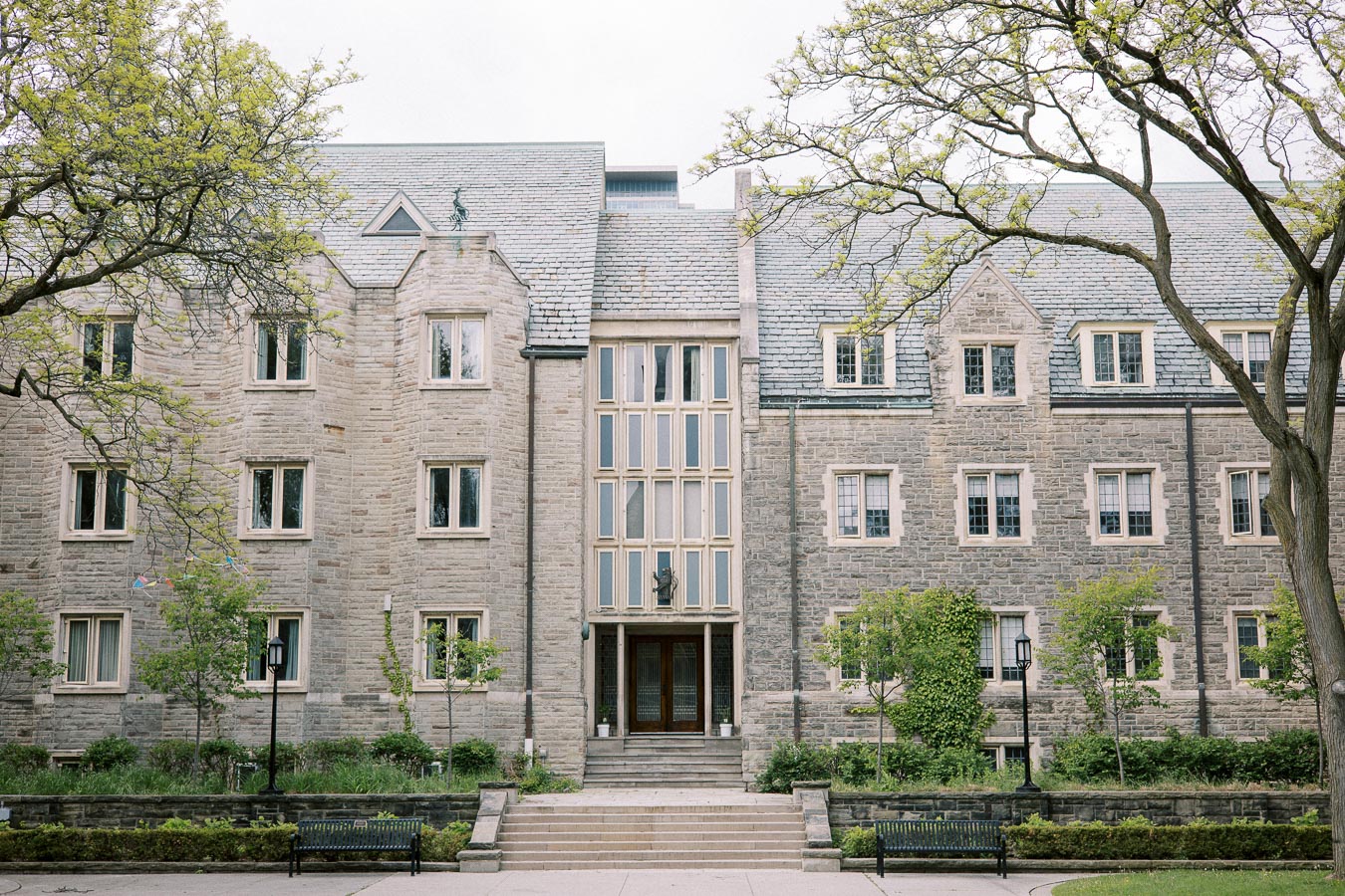 Historic stone building with numerous windows, surrounded by trees and greenery, featuring central entrance steps and benches in an academic setting.