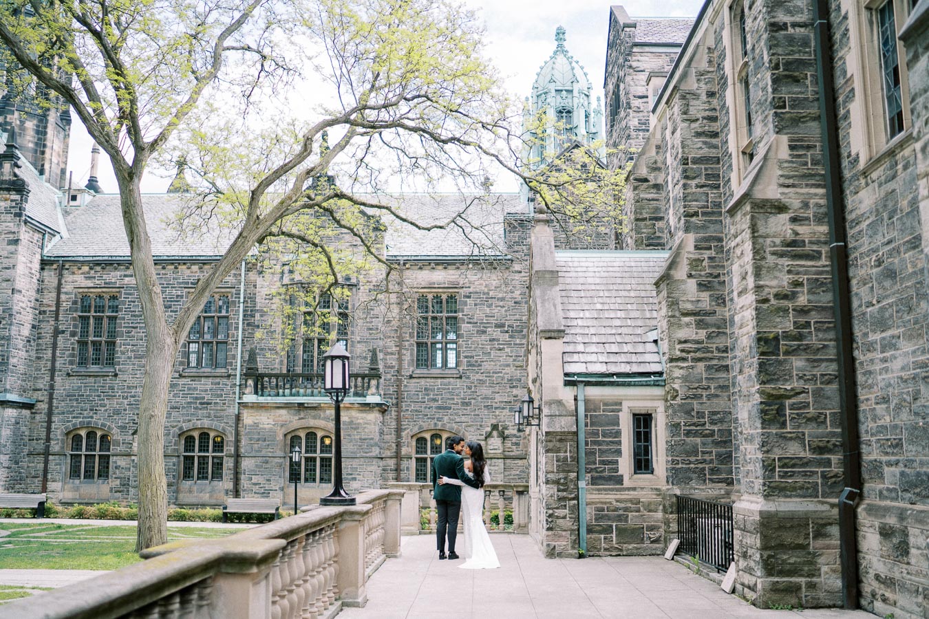 Romantic couple embracing in front of historic stone building with arched windows, surrounded by lush greenery on a serene pathway.
