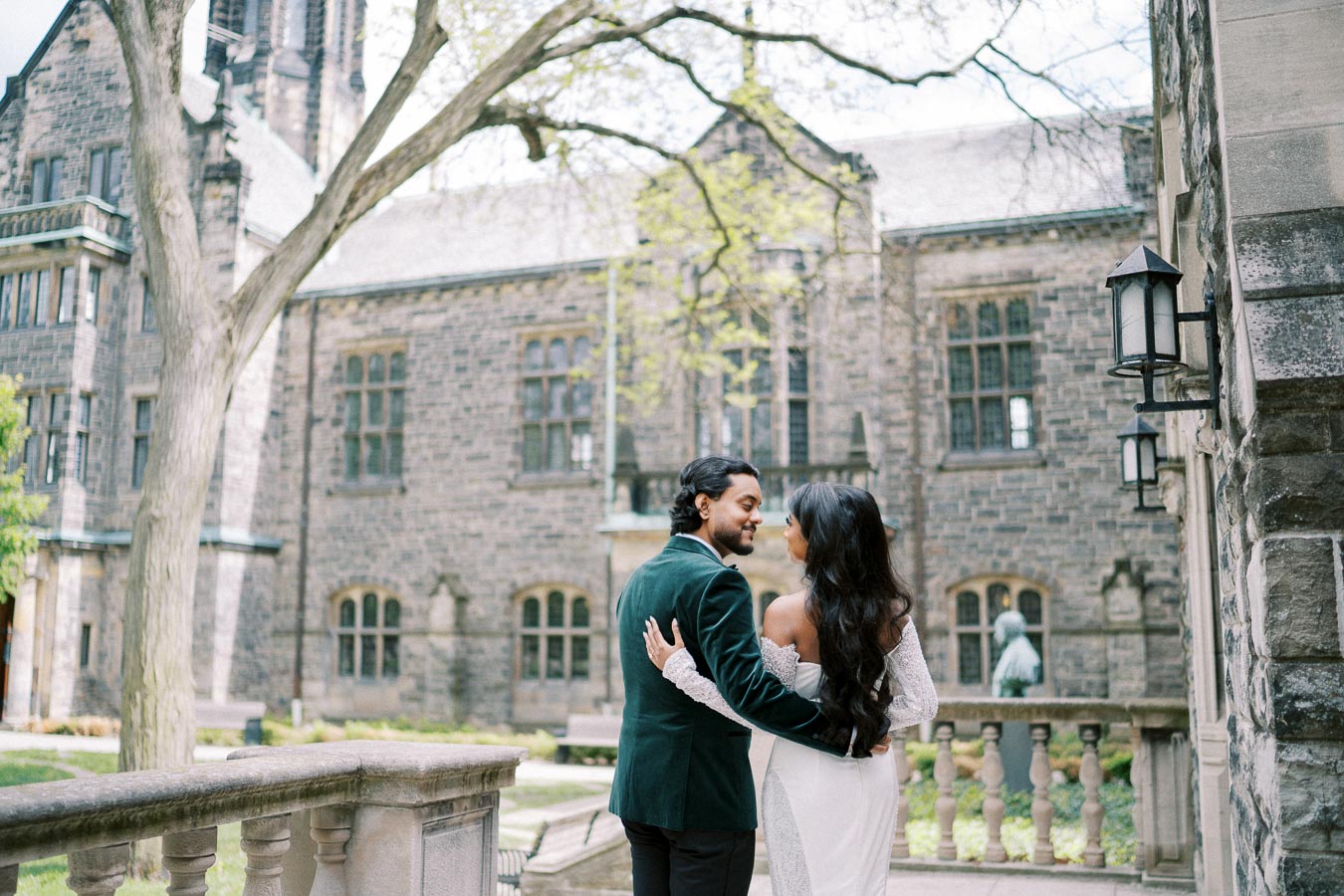 A couple embracing in a romantic moment outside a historic stone building with Gothic architecture, surrounded by greenery and an ornate railing, on a bright day.