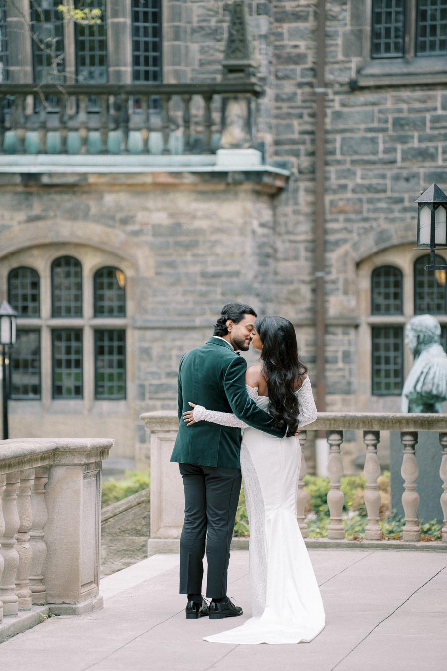 A couple dressed elegantly in formal attire shares a romantic moment near a historic stone building with large windows.