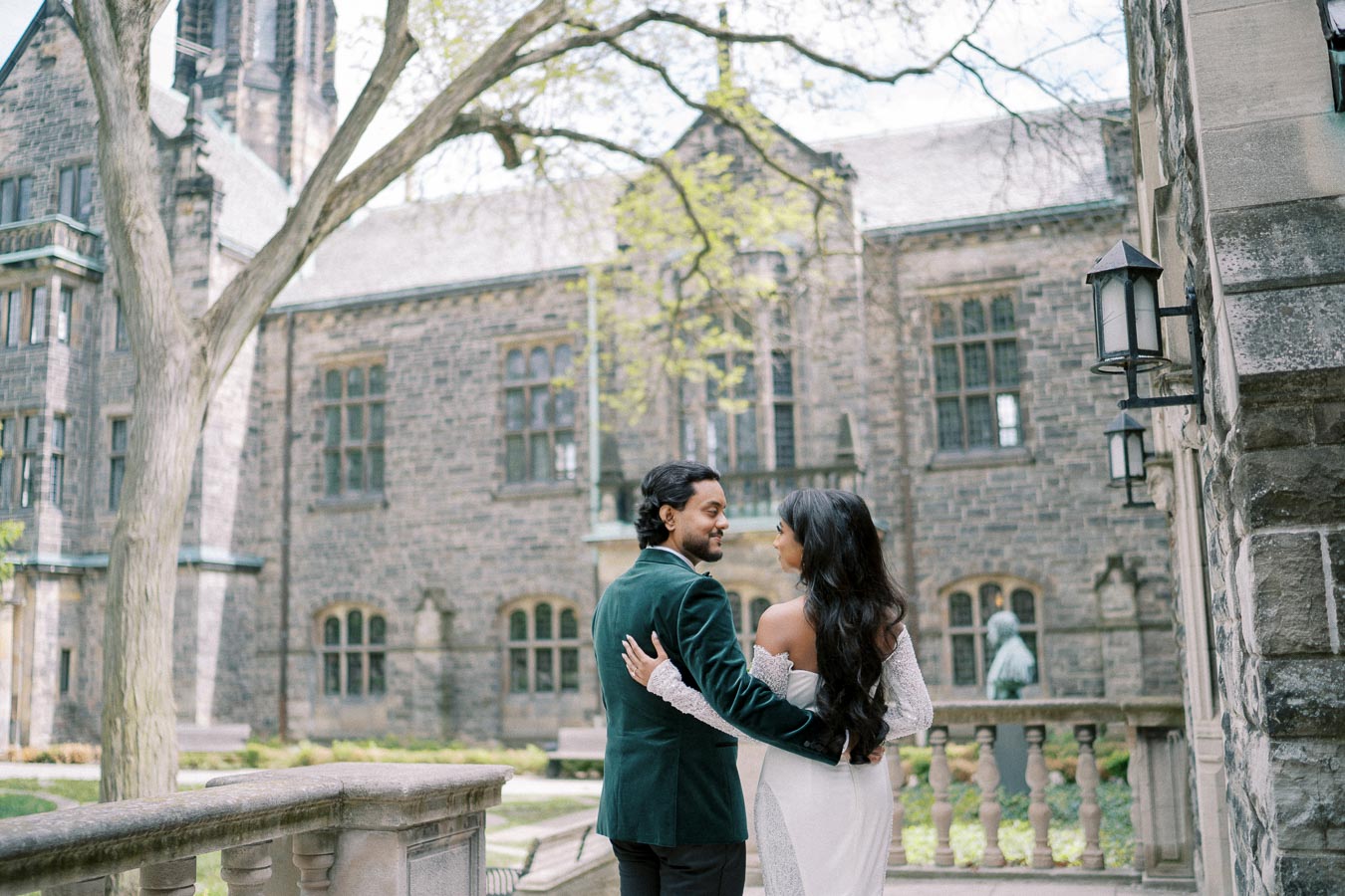A couple embracing in an elegant outdoor setting with historic stone architecture in the background, under a large tree on a sunny day.