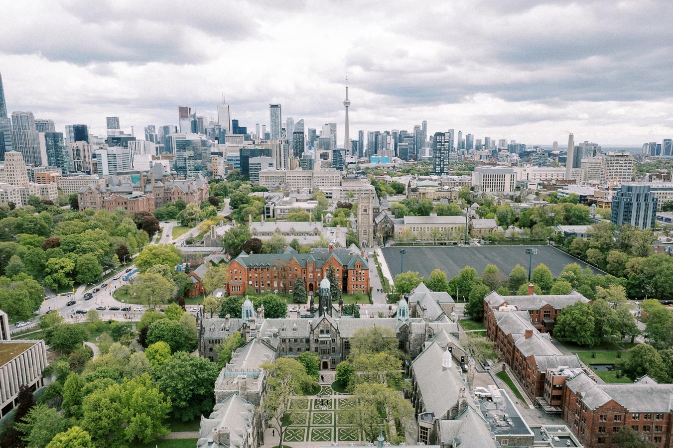 Aerial view of downtown Toronto featuring the University of Toronto campus, historic buildings, lush greenery, and the iconic CN Tower in the background under a cloudy sky.