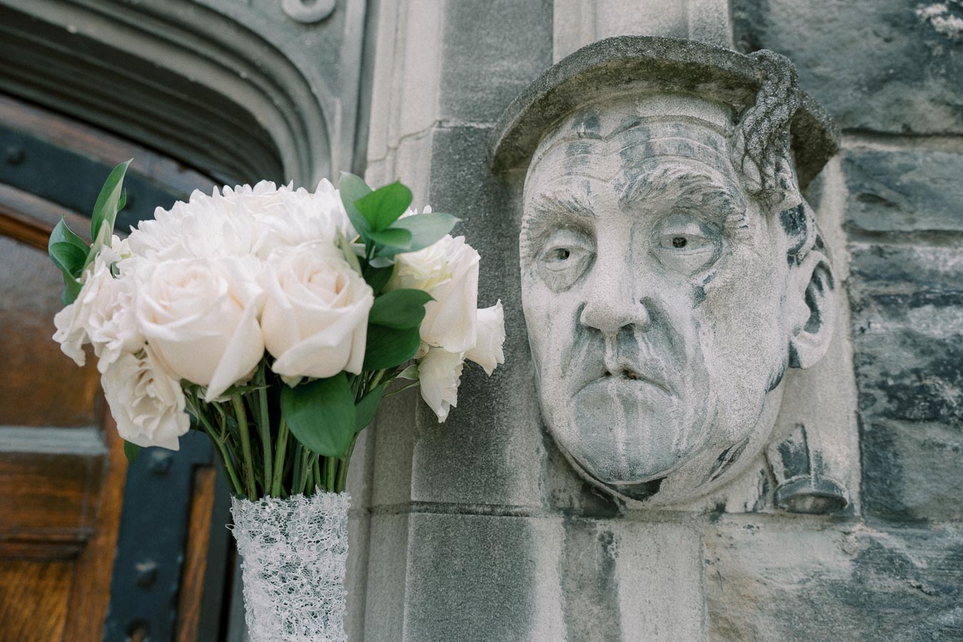 Stone sculpture of a solemn face on a historic building facade with a bouquet of white roses in the foreground