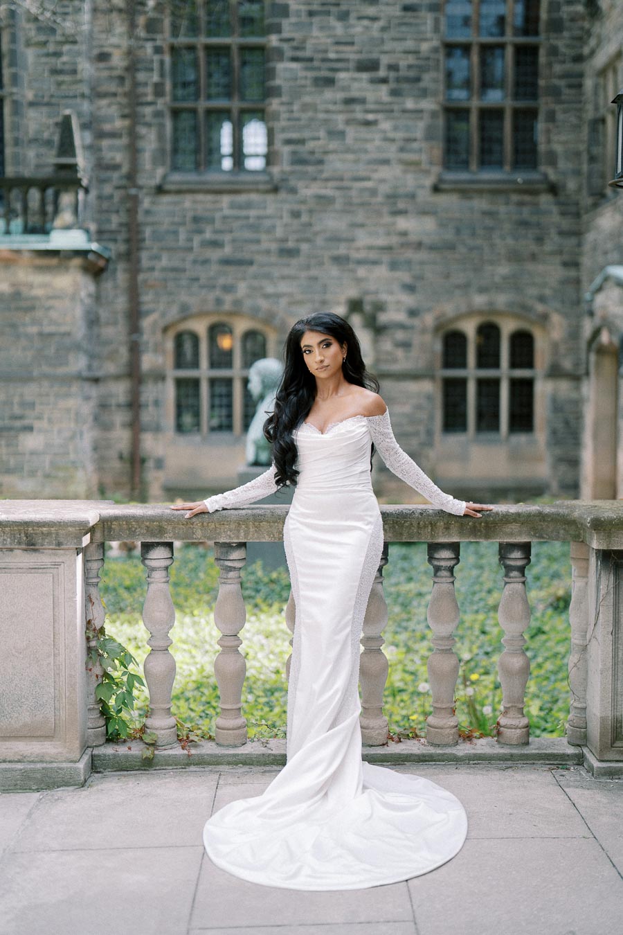 A woman in a beautiful, off-the-shoulder white gown poses elegantly on a stone balcony in front of a historic stone building. Her long, dark hair cascades over her shoulders, complementing the sophisticated bridal look. Elegant wedding fashion.