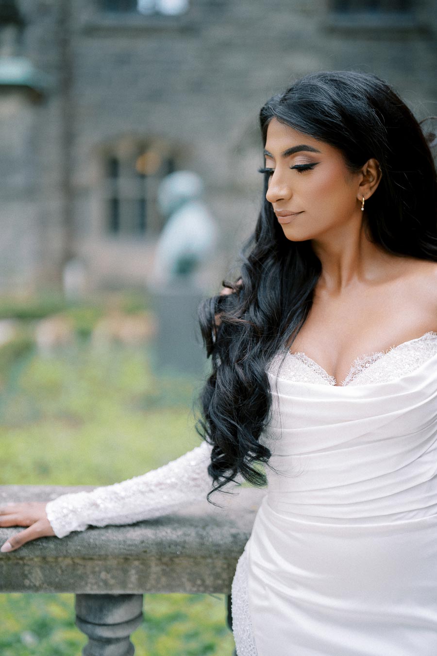 Elegant bride in a white wedding dress with lace details, posing outdoors against a stone railing and greenery.