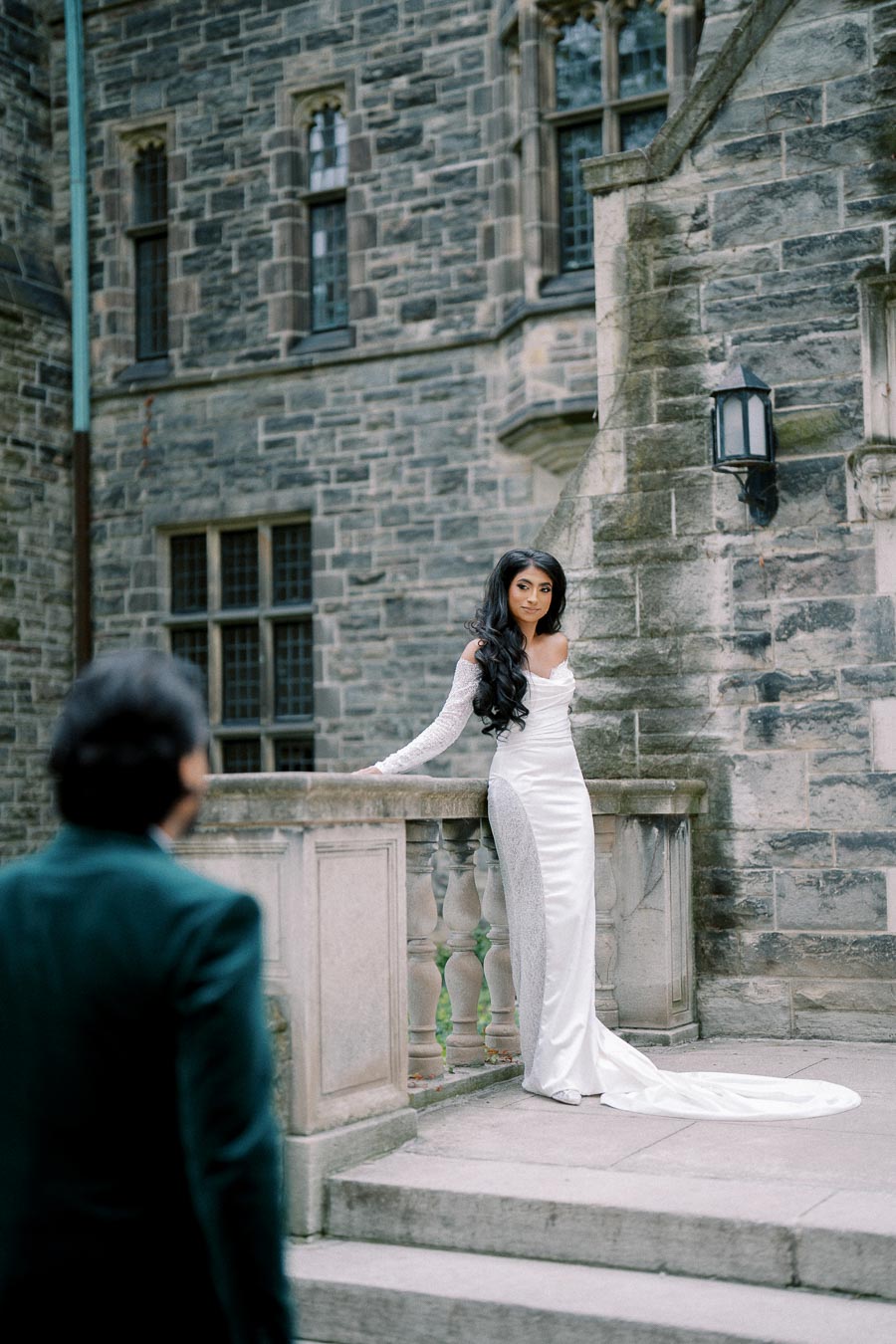 Woman in elegant white gown standing on stone staircase at historic building, with person approaching, capturing a romantic and timeless moment.