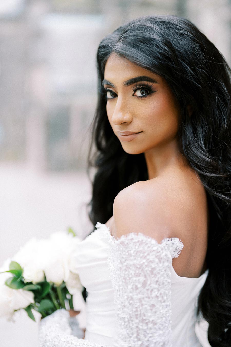 Bridal portrait of a woman with long black hair holding a bouquet of white flowers, wearing a white dress with lace details, captured in soft natural lighting.