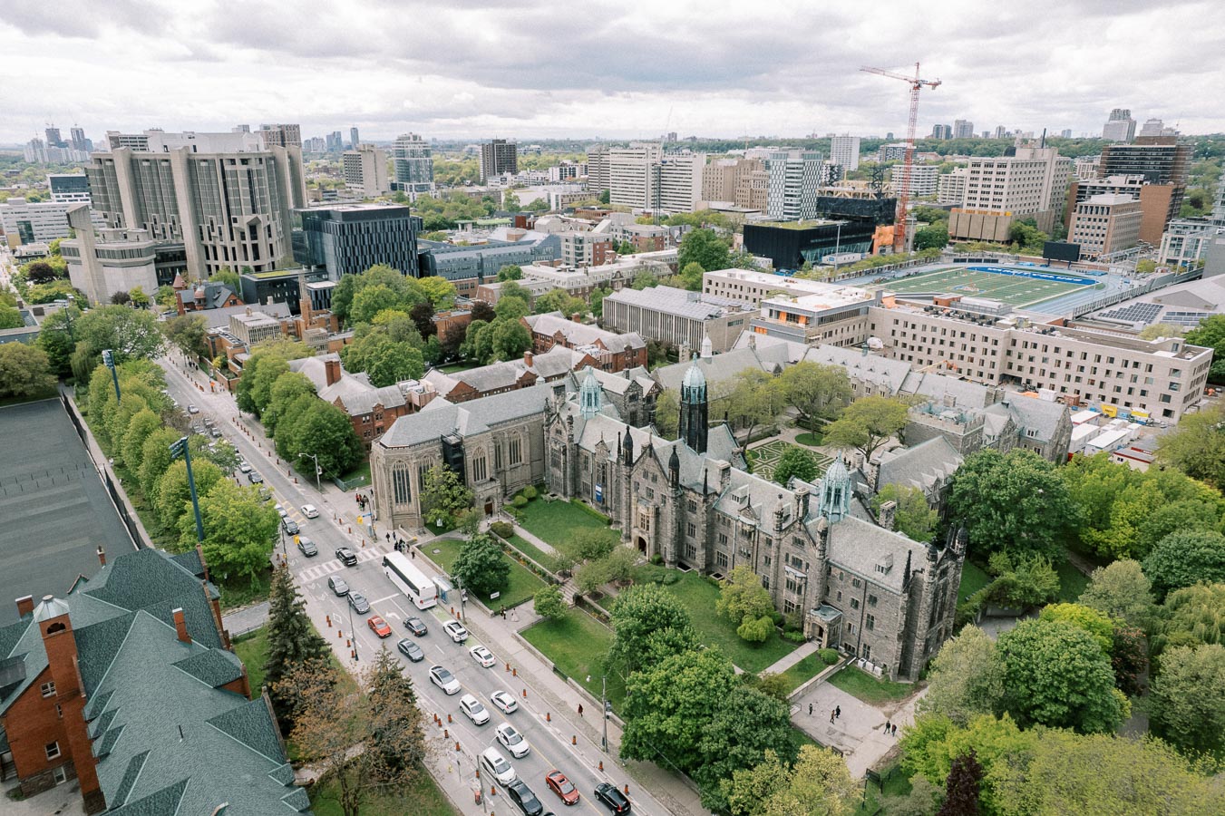 Aerial view of a university campus featuring Gothic-style architecture, modern buildings, lush green spaces, and a sports field, set against an urban skyline under a cloudy sky.