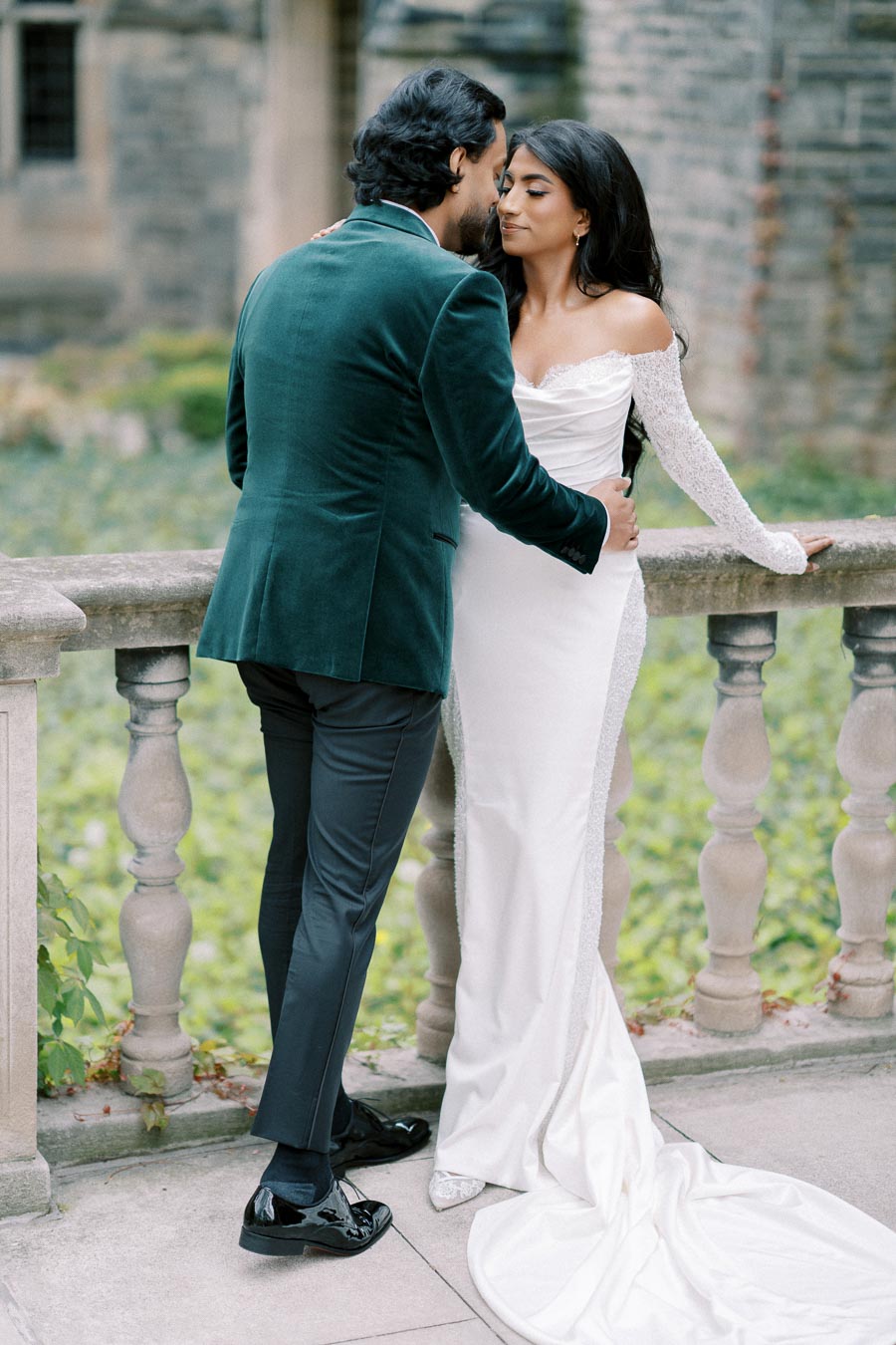 A groom in a dark green velvet jacket embraces his bride in a white off-shoulder wedding gown beside a historic stone balustrade, surrounded by natural greenery, capturing a romantic moment.